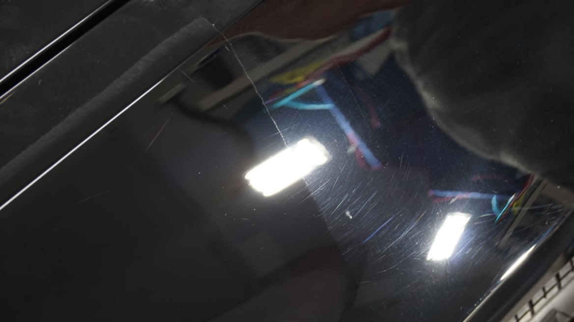 Błyszcząca, czarna karoseria samochodu z widocznymi rysami pod światło lampy warsztatowej, ukazująca stan lakieru przed polerowaniem lub renowacją.