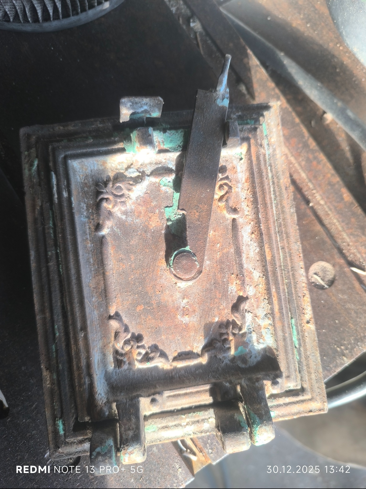 Zardzewiała, kwadratowa klapka z metalu z dekoracyjnymi elementami i zielonymi śladami korozji, uchwyt w pozycji otwartej, leżąca na metalowej powierzchni.