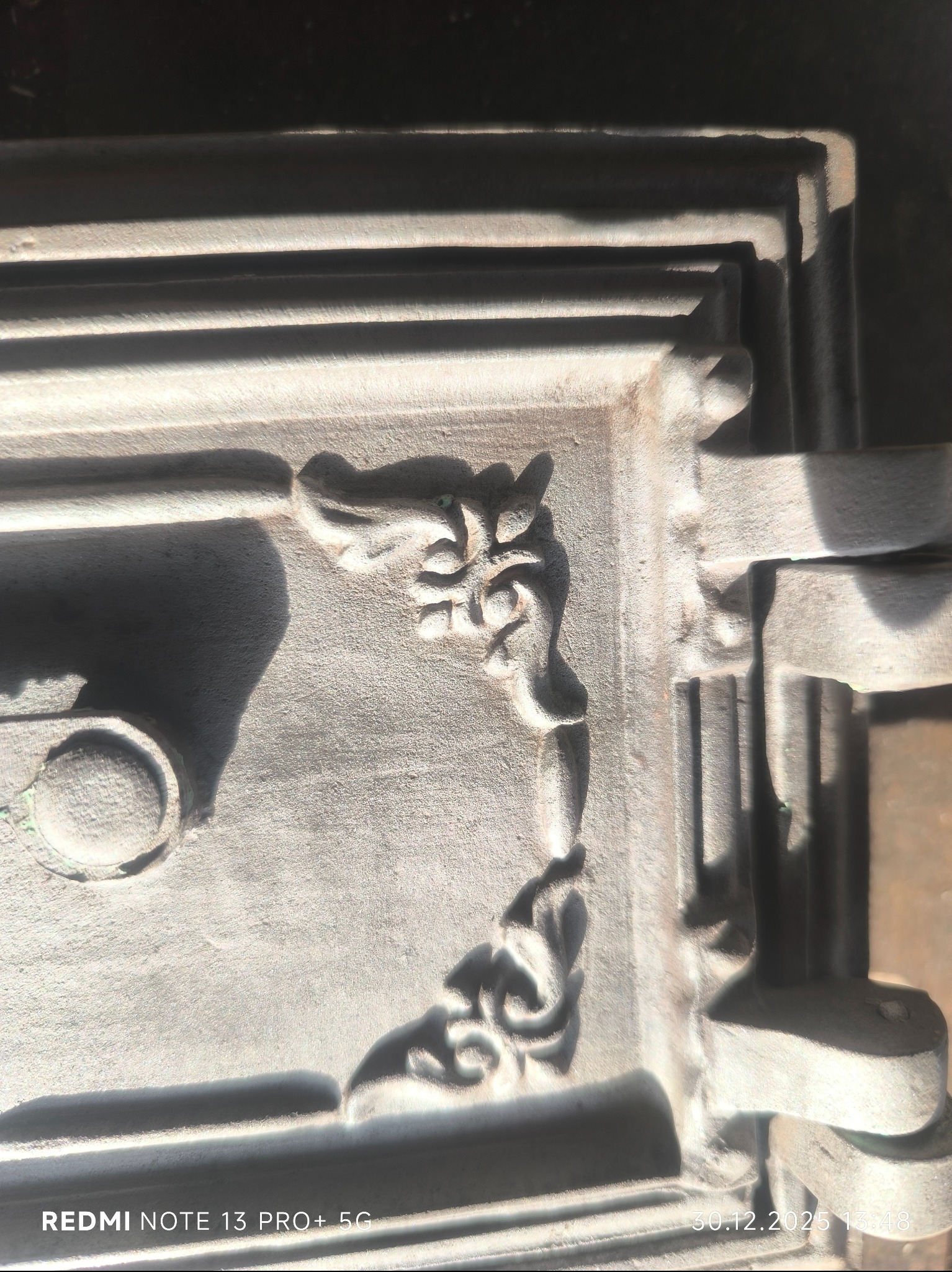 Detal metalowej konstrukcji z ornamentami, oświetlonej z góry, ukazującej fakturę i cieniowanie. Widoczne zdobienia w narożniku i fragmenty konstrukcji.