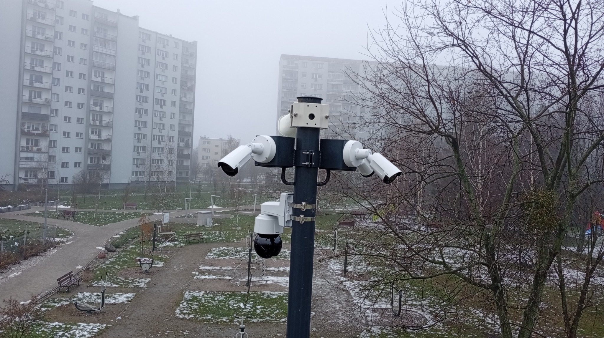 Zestaw kamer monitoringu na słupie, skierowanych w różne strony, z widokiem na osiedle w mglisty dzień. Kamery białe, słup szary.