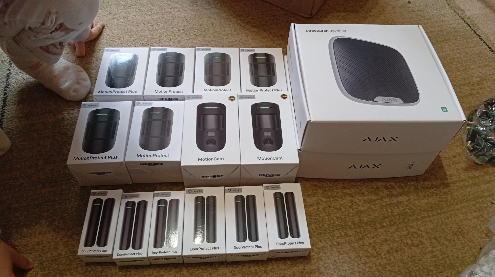 Zestaw komponentów systemu alarmowego Ajax w oryginalnych opakowaniach: czujniki ruchu, otwarcia drzwi i syrena alarmowa. Widoczne modele MotionProtect, DoorProtect i StreetSiren.