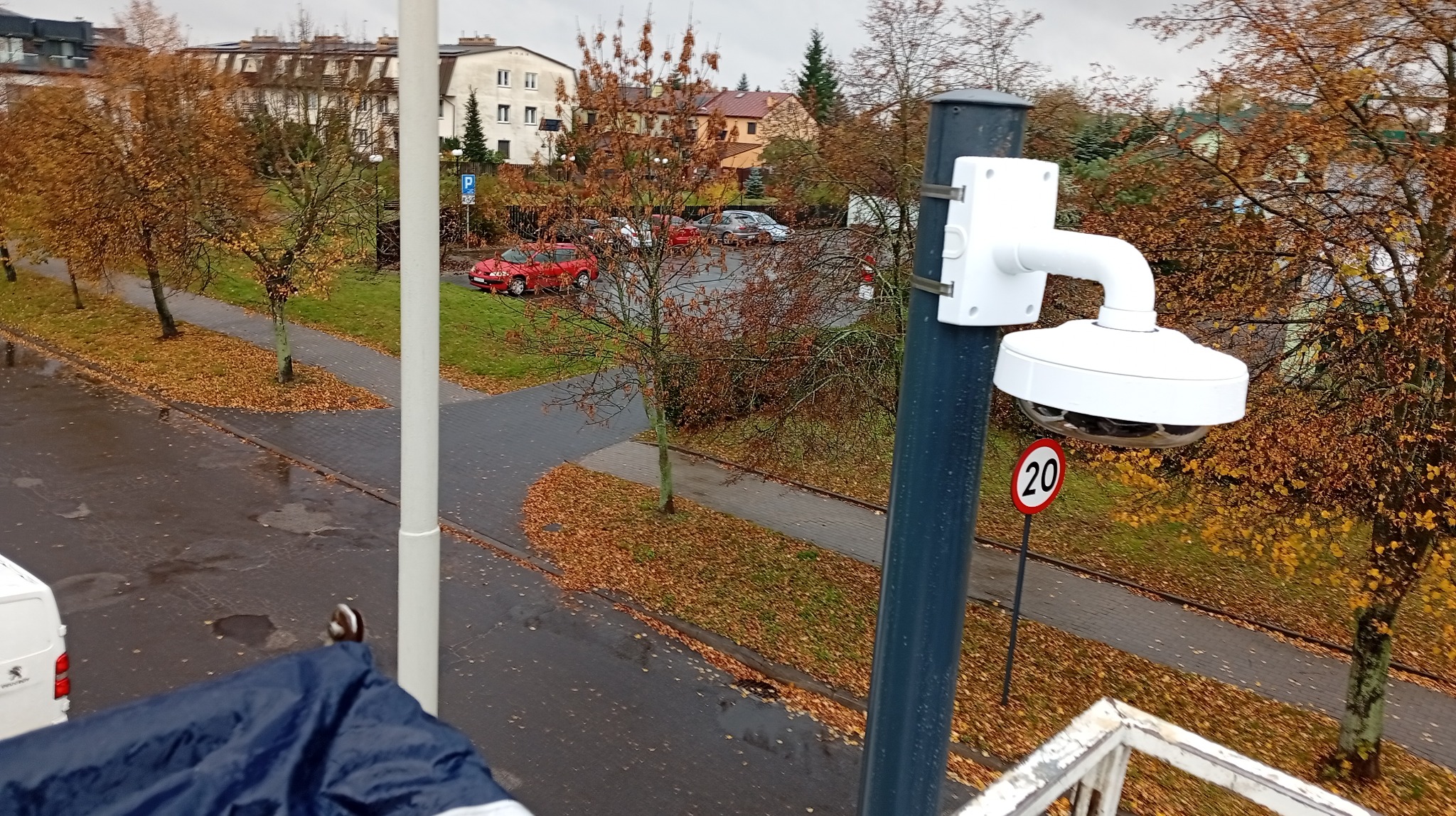 Kamera monitoringu typu rybie oko zamontowana na słupie, widok z góry na parking w jesiennej scenerii. Widoczny znak ograniczenia prędkości do 20 km/h.