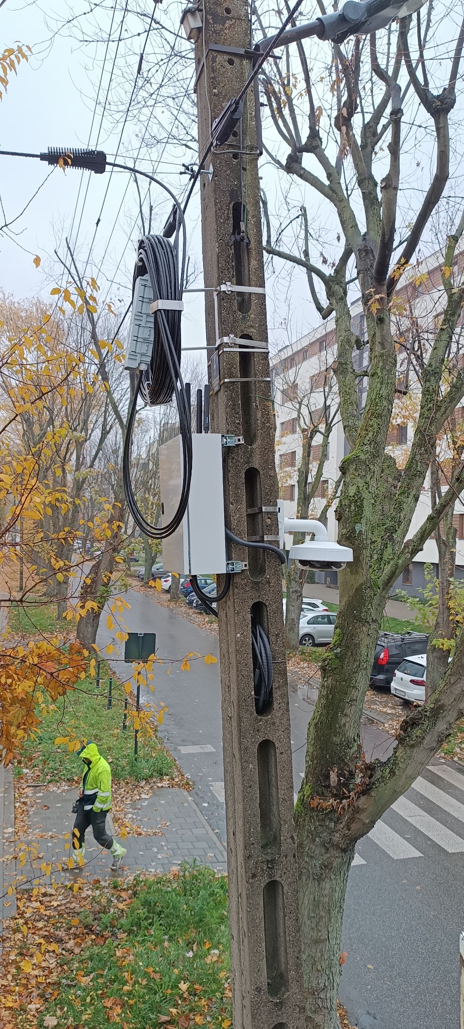 Kamera monitoringu zamontowana na słupie energetycznym, widok z góry na ulicę z przechodniem w odblaskowej kurtce. Jesienne drzewa w tle.