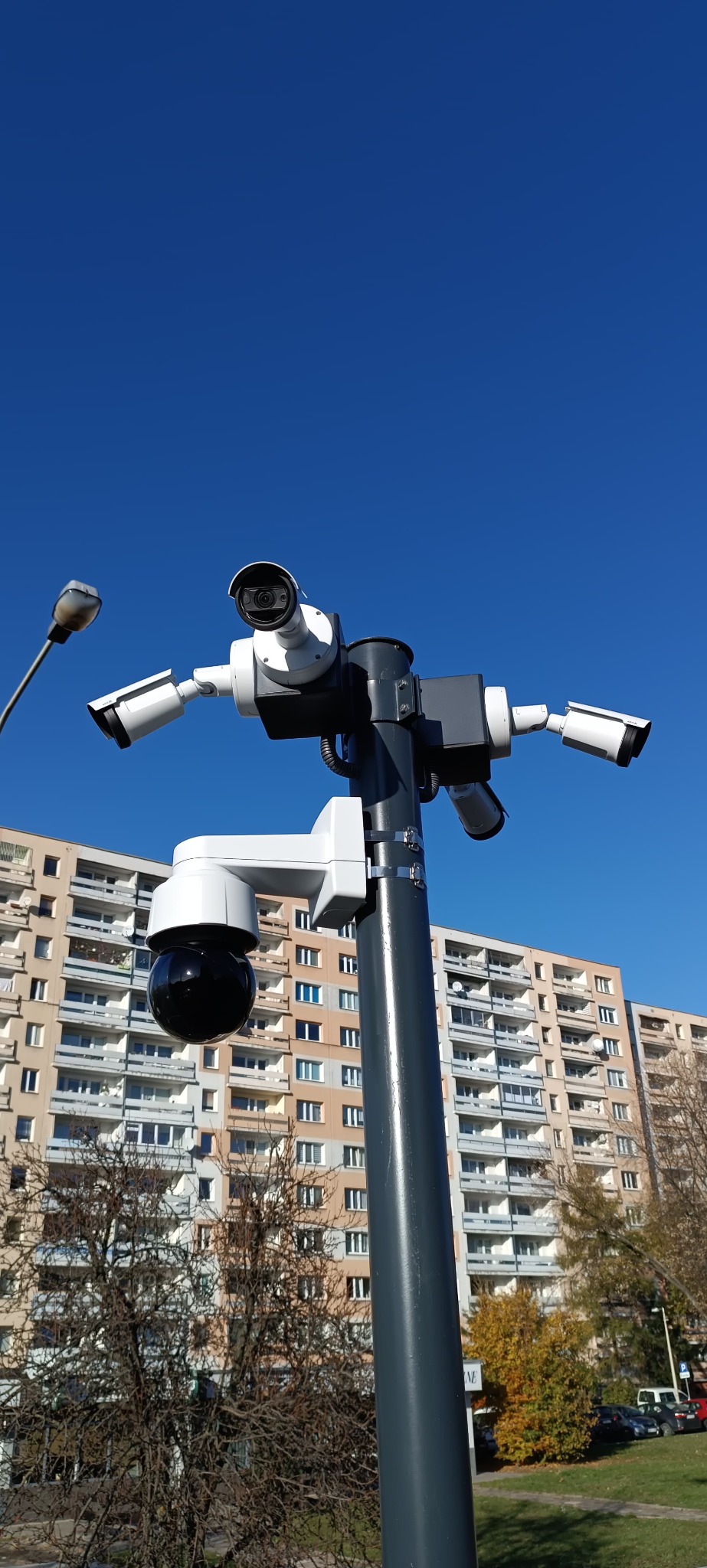 Słup z zestawem kamer monitoringu (PTZ i tubowe) na tle bloku mieszkalnego i błękitnego nieba. Widok z dołu, podkreślający rozbudowany system zabezpieczeń.