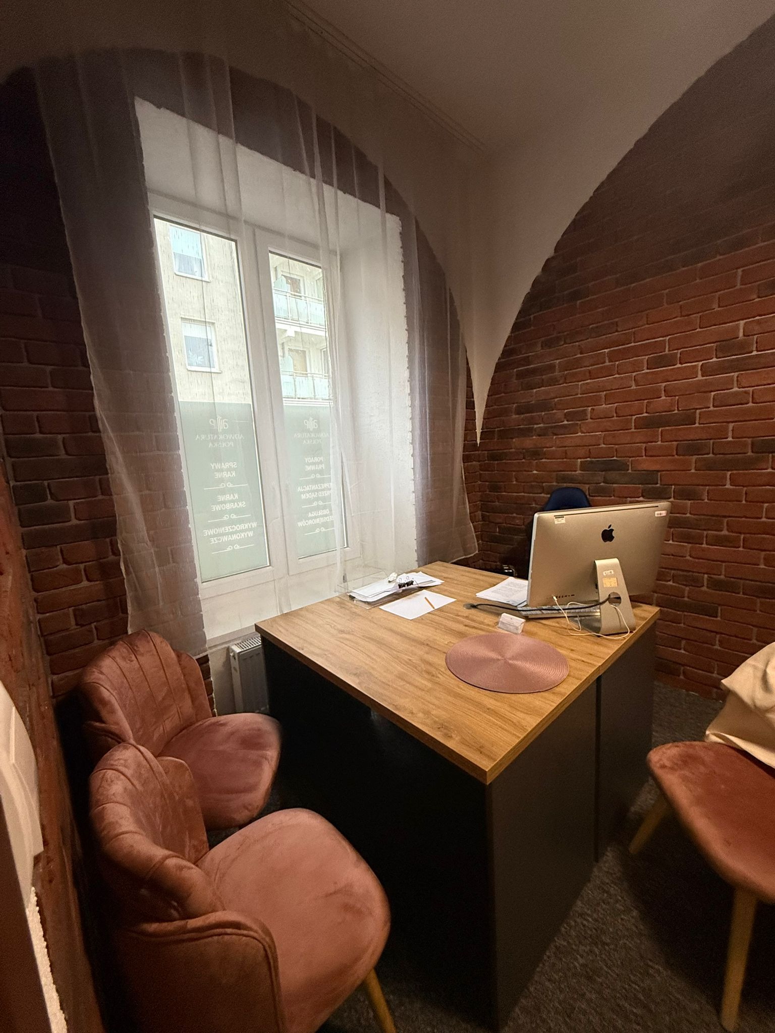 Biuro z ceglaną ścianą, drewnianym biurkiem, komputerem iMac i krzesłami w odcieniach różu. Okno z widokiem na budynek. Nowoczesne wnętrze kancelarii.
