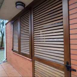 Tokarski Jacek - Drewniane okiennice i drzwi lamelowe w brązowej ramie, zamontowane na ceglanej ścianie budynku z widocznym oświetleniem zewnętrznym i fragmentem otoczenia.