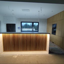 Tokarski Jacek - Nowoczesna wyspa kuchenna z drewnianymi lamelami i podświetleniem LED, w tle zabudowa kuchenna z forniru i okno z widokiem na zewnątrz. Wnętrze w trakcie wykończenia.