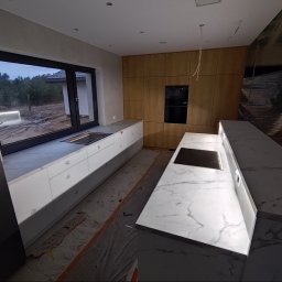 Tokarski Jacek - Nowoczesna kuchnia z białymi szafkami i marmurowym blatem, duże okno z widokiem na zewnątrz. Widoczne drewniane panele na ścianie i płyta indukcyjna w blacie.