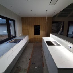 Tokarski Jacek - Montaż kuchni na wymiar: białe blaty z marmurowym wzorem, drewniane szafki, okno z widokiem na zewnątrz. Widoczne elementy instalacji, etap prac wykończeniowych.