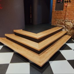 Tokarski Jacek - Trójkątne, drewniane schody z czarnymi wstawkami na czarno-białej podłodze w szachownicę, obok ceglanej ściany z elementami kamiennymi i czarnym znakiem toalety.