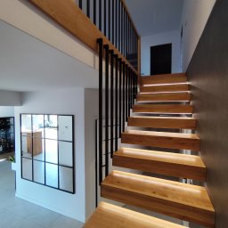 Tokarski Jacek - Nowoczesne schody drewniane z podświetleniem LED, czarne metalowe barierki, minimalistyczny design, jasne wnętrze z lustrem w czarnej ramie.