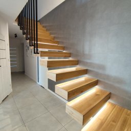Tokarski Jacek - Nowoczesne schody drewniane wiszące z podświetleniem LED, czarne metalowe barierki, szara ściana z płytek, jasna podłoga. Minimalistyczny design.