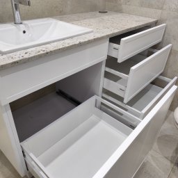 Tokarski Jacek - Biała szafka łazienkowa z umywalką i blatem z jasnego granitu, trzy wysuwane szuflady z systemem Blum, widoczna toaleta i płytki w tle. Ujęcie z bliska.