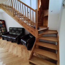Tokarski Jacek - Drewniane schody z balustradą w jasnym wnętrzu, obok ciemna skórzana sofa i regał z książkami. Parkiet ułożony w jodełkę. Widoczne drugie schody w tle.