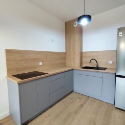 Tokarski Jacek - Szara zabudowa kuchenna z drewnianym blatem i panelem ściennym, czarny zlew i płyta indukcyjna, nowoczesna lampa sufitowa, jasna podłoga. Minimalistyczny design.