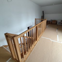 Tokarski Jacek - Drewniana balustrada schodowa z jasnego drewna, widok z góry, zabezpieczona podłoga kartonem, białe ściany, surowe wykończenie, wnętrze domu w trakcie prac.