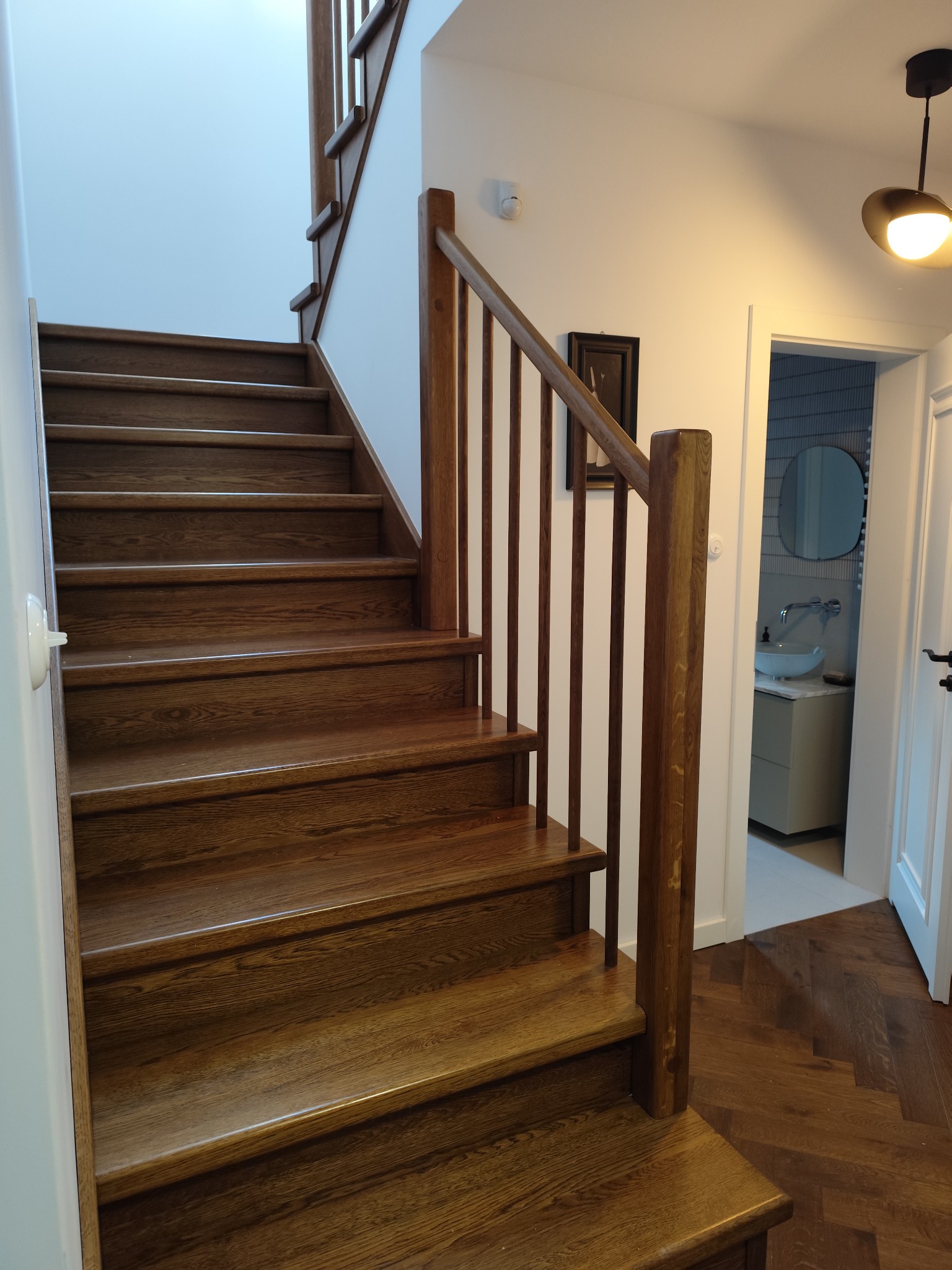 Drewniane schody z balustradą w Aleksandrowie Kujawskim, prowadzące na piętro. Widoczna podłoga ułożona w jodełkę i wejście do łazienki z umywalką.