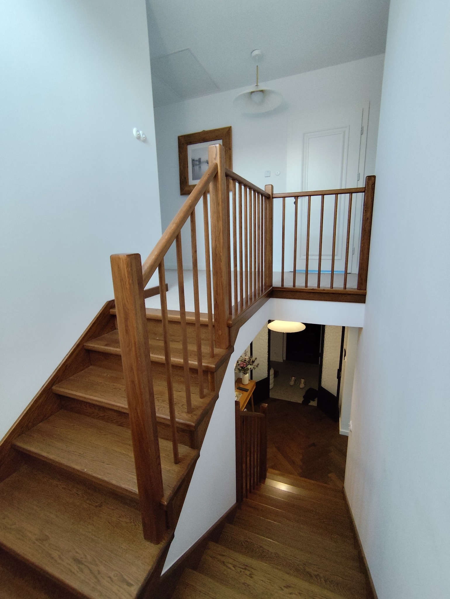 Drewniane schody z balustradą w jasnym wnętrzu. Widok z góry na dół, ukazujący naturalne wykończenie drewna i minimalistyczny design. W tle widoczny fragment korytarza.