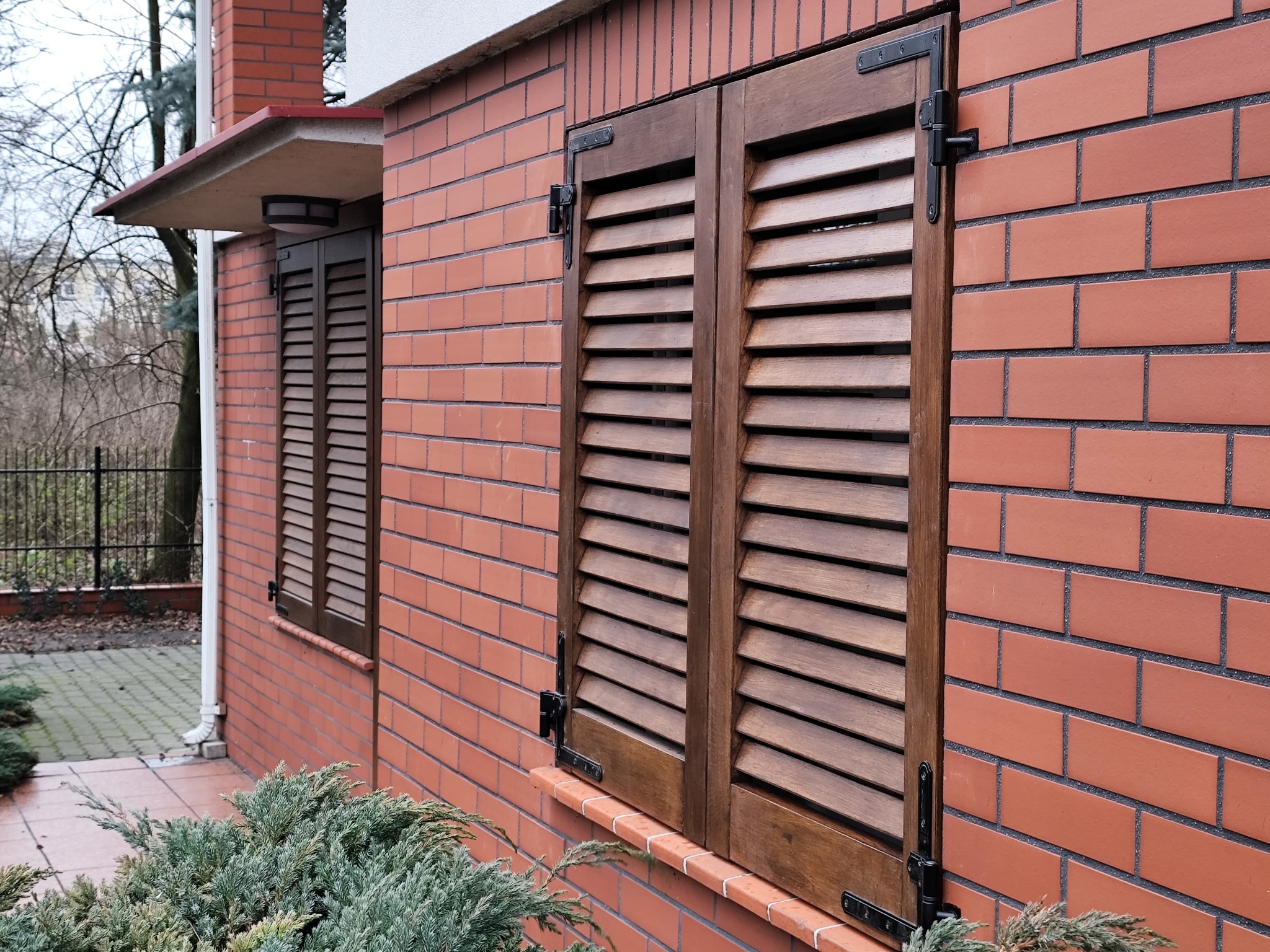 Drewniane okiennice z żaluzjami, zamontowane na ceglanej ścianie budynku. Widoczne czarne okucia i fragment roślinności u dołu kadru. Ujęcie z dołu.
