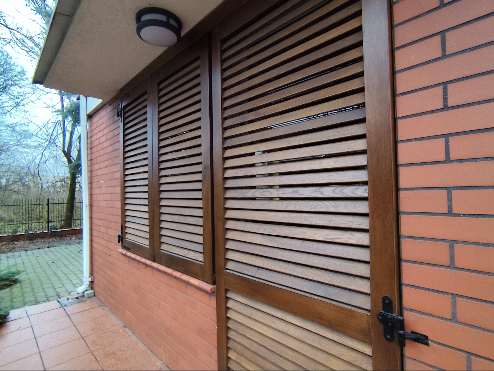Drewniane okiennice i drzwi lamelowe w brązowej ramie, zamontowane na ceglanej ścianie budynku z widocznym oświetleniem zewnętrznym i fragmentem otoczenia.