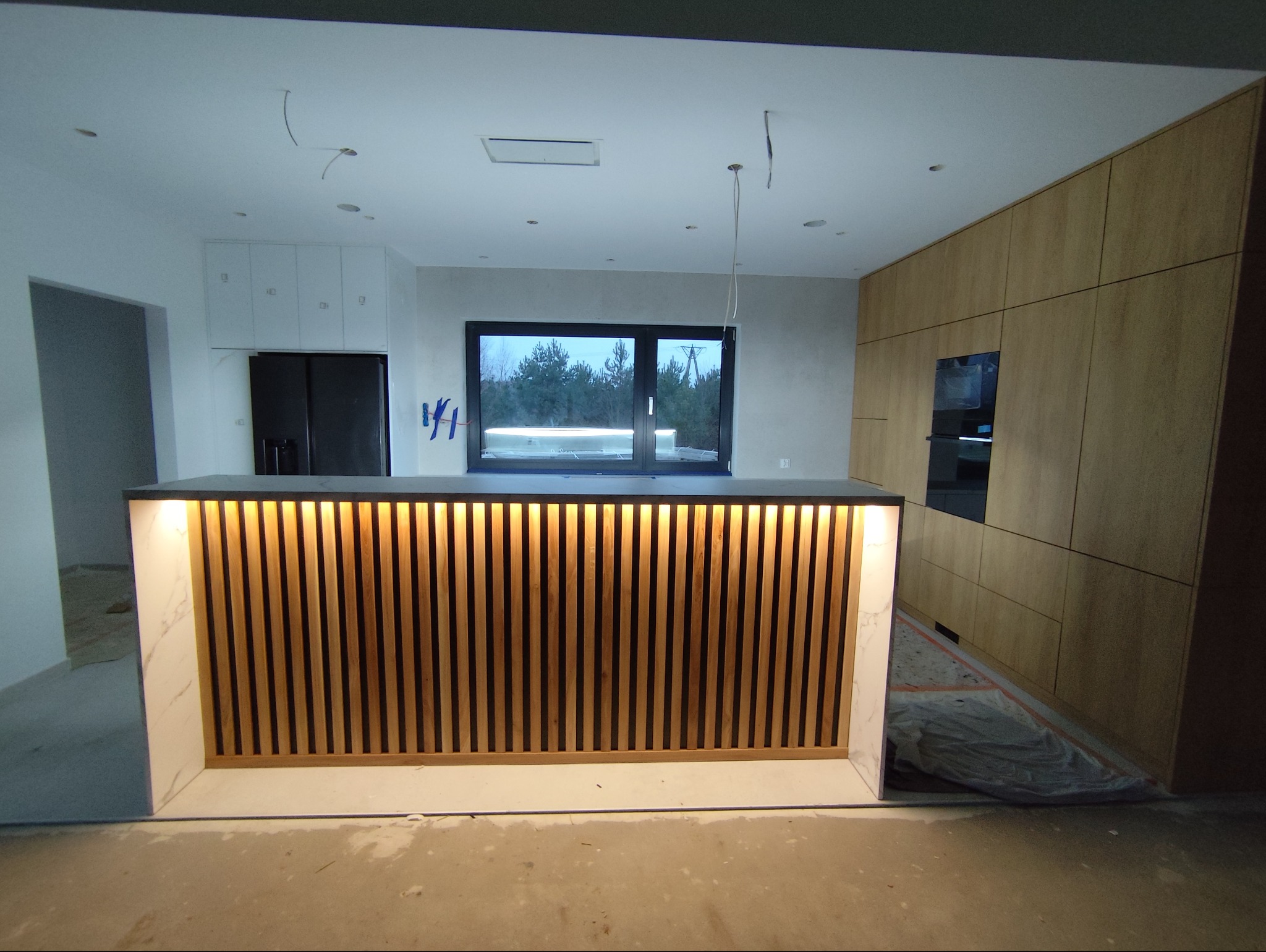 Nowoczesna wyspa kuchenna z drewnianymi lamelami i podświetleniem LED, w tle zabudowa kuchenna z forniru i okno z widokiem na zewnątrz. Wnętrze w trakcie wykończenia.