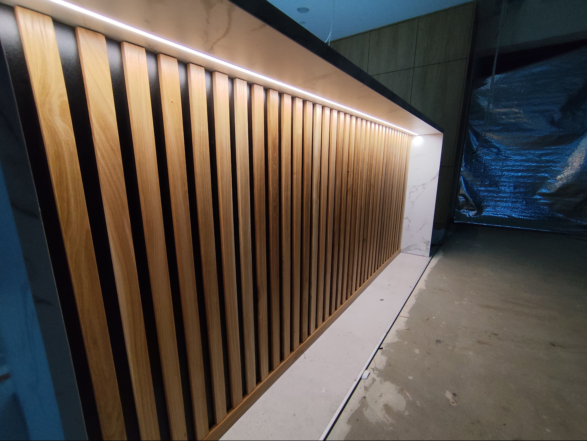 Nowoczesna zabudowa z drewnianych lameli i podświetleniem LED, wykończona marmurem. Widoczny fragment podłogi w trakcie prac wykończeniowych.