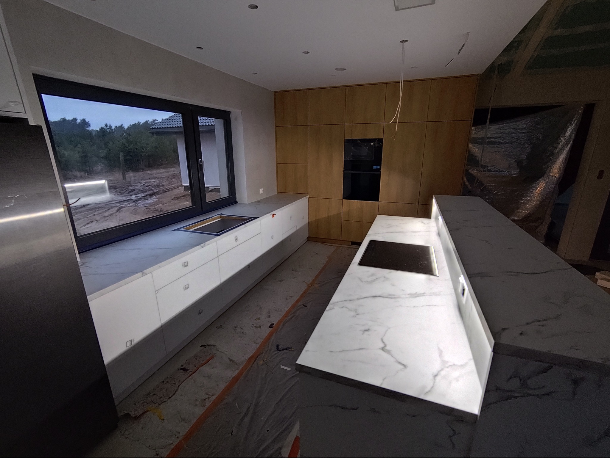 Nowoczesna kuchnia z białymi szafkami i marmurowym blatem, duże okno z widokiem na zewnątrz. Widoczne drewniane panele na ścianie i płyta indukcyjna w blacie.