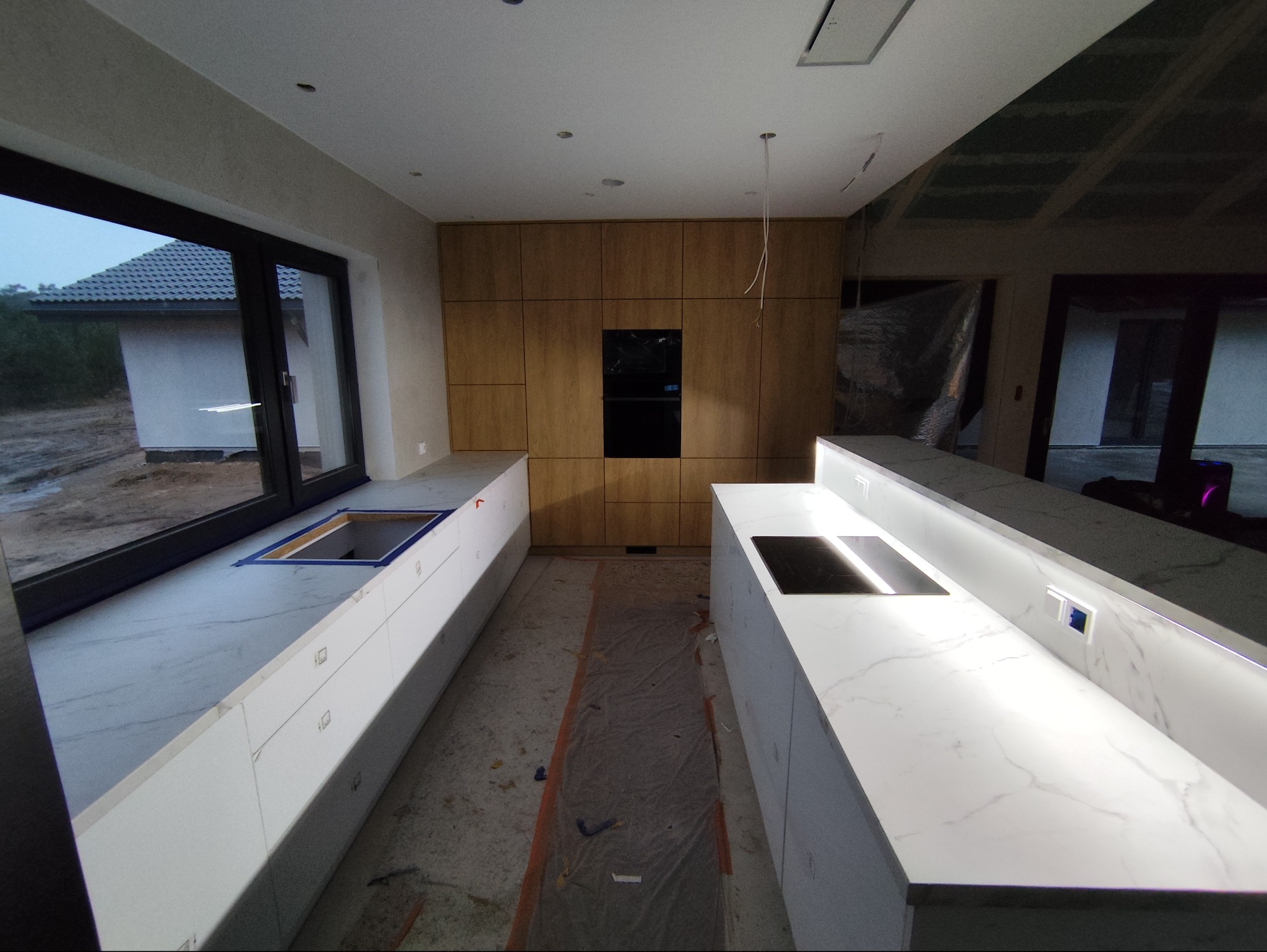 Montaż kuchni na wymiar: białe blaty z marmurowym wzorem, drewniane szafki, okno z widokiem na zewnątrz. Widoczne elementy instalacji, etap prac wykończeniowych.