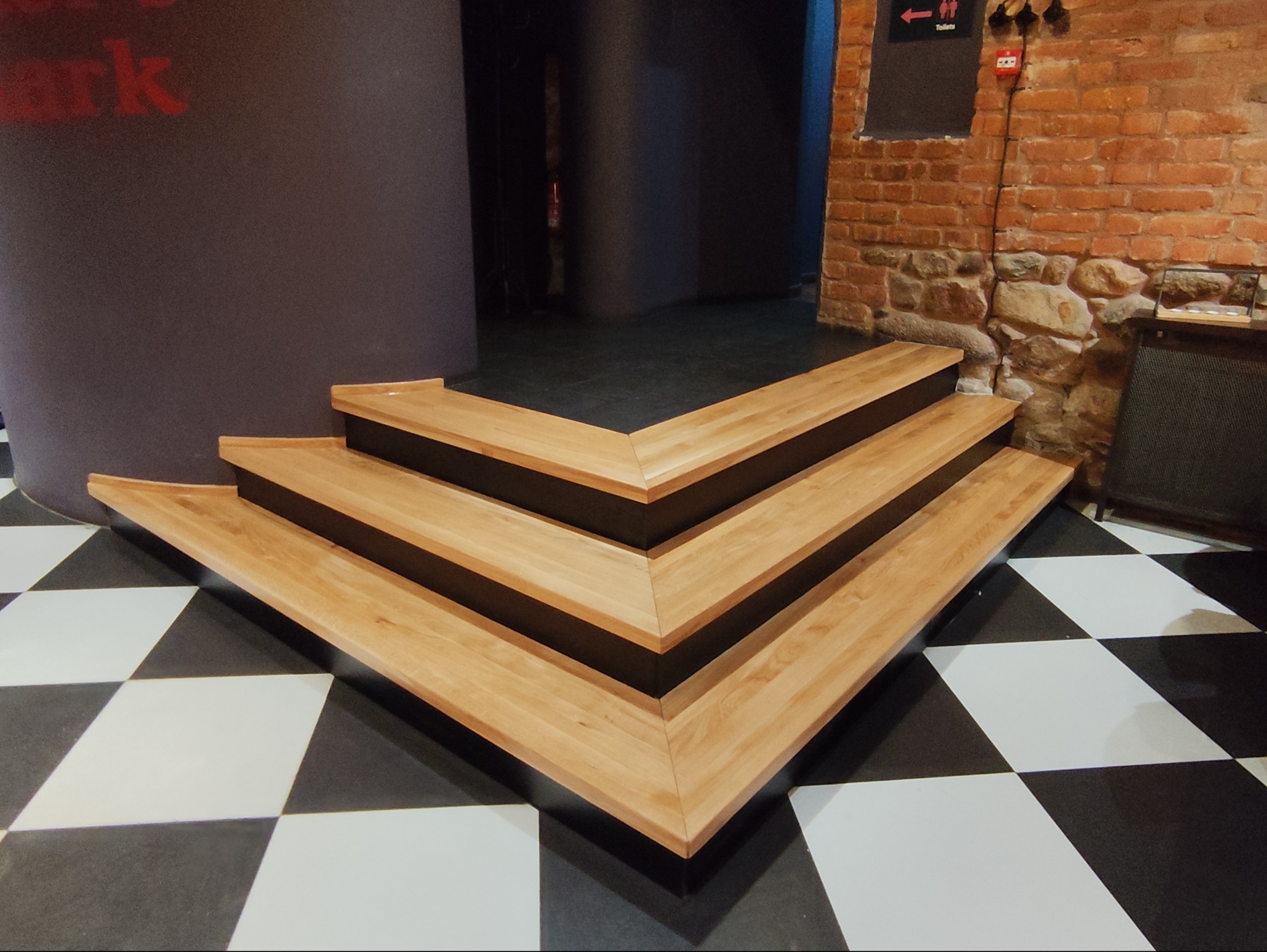 Trójkątne, drewniane schody z czarnymi wstawkami na czarno-białej podłodze w szachownicę, obok ceglanej ściany z elementami kamiennymi i czarnym znakiem toalety.