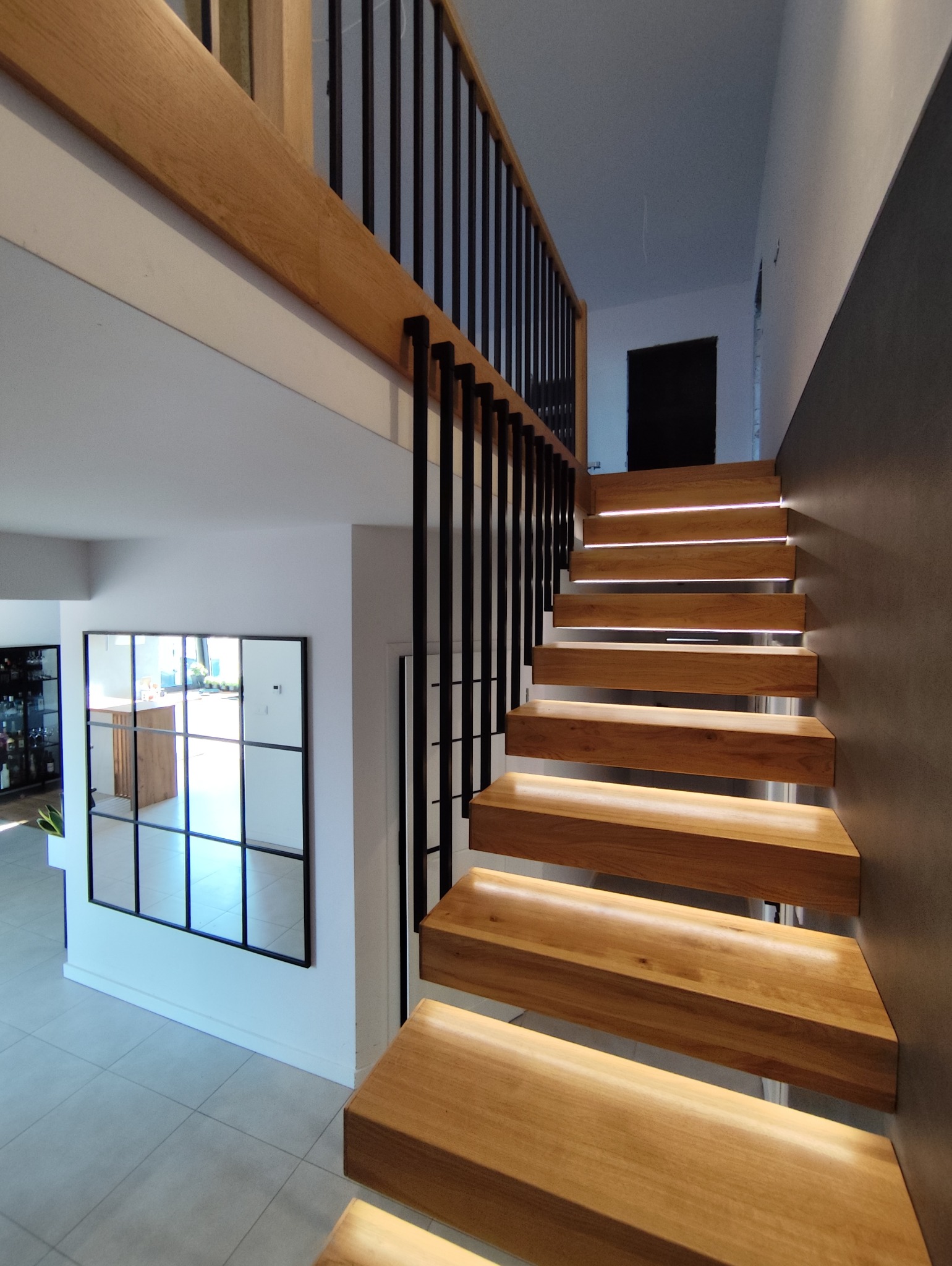 Nowoczesne schody drewniane z podświetleniem LED, czarne metalowe barierki, minimalistyczny design, jasne wnętrze z lustrem w czarnej ramie.