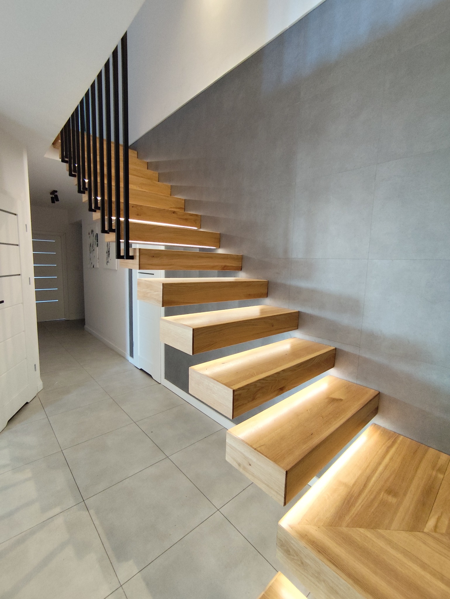 Nowoczesne schody drewniane wiszące z podświetleniem LED, czarne metalowe barierki, szara ściana z płytek, jasna podłoga. Minimalistyczny design.