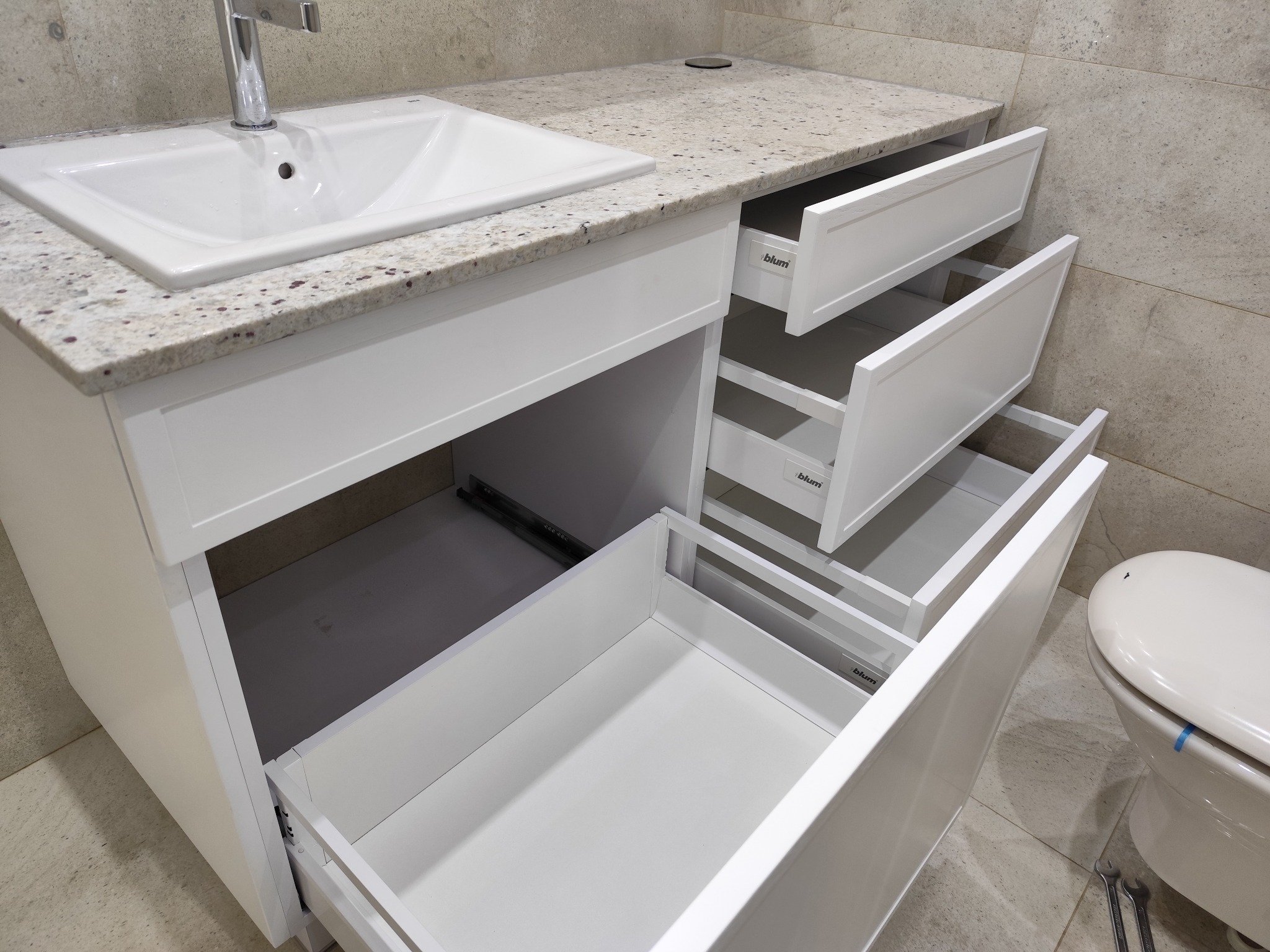 Biała szafka łazienkowa z umywalką i blatem z jasnego granitu, trzy wysuwane szuflady z systemem Blum, widoczna toaleta i płytki w tle. Ujęcie z bliska.