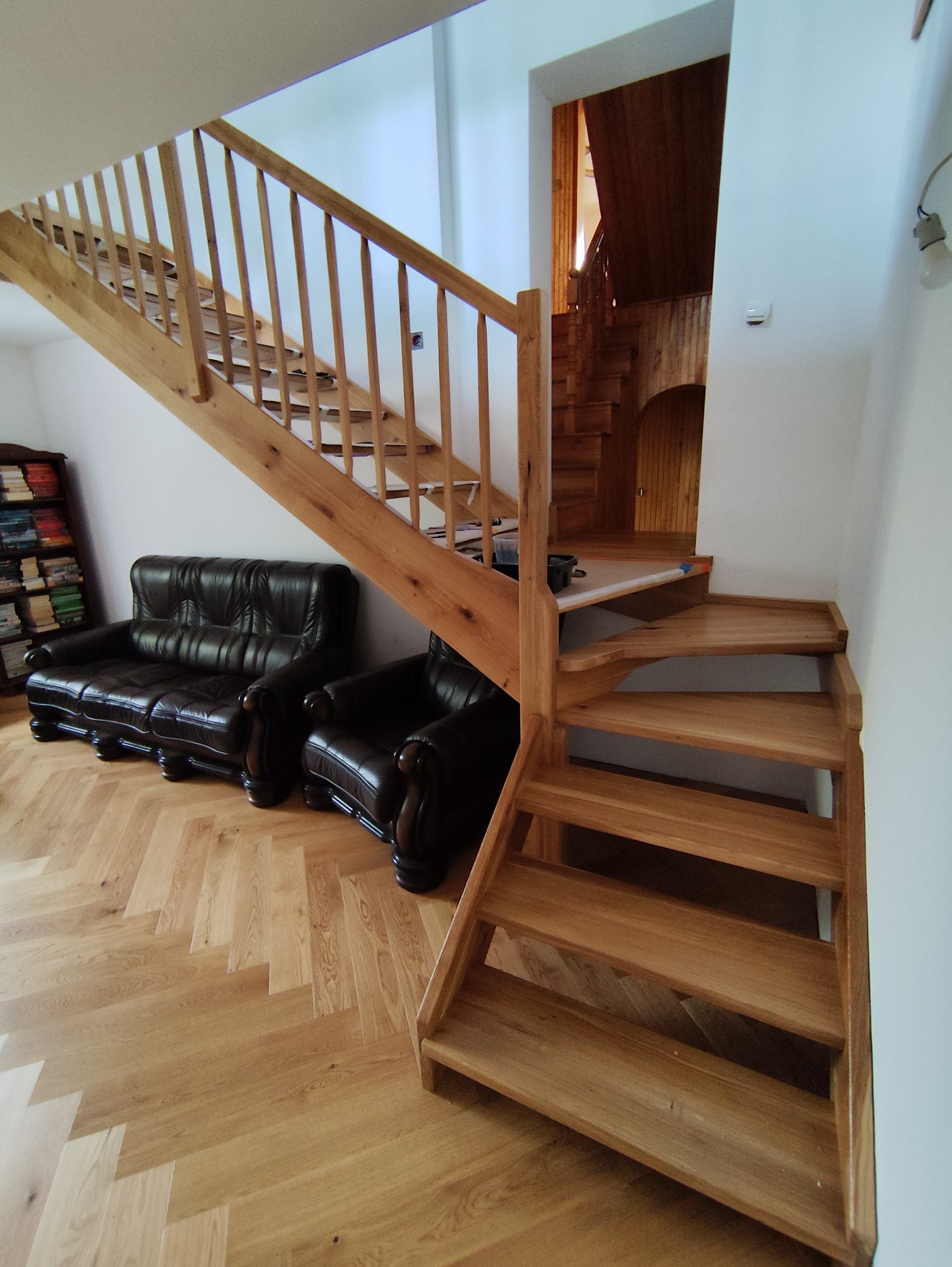 Drewniane schody z balustradą w jasnym wnętrzu, obok ciemna skórzana sofa i regał z książkami. Parkiet ułożony w jodełkę. Widoczne drugie schody w tle.