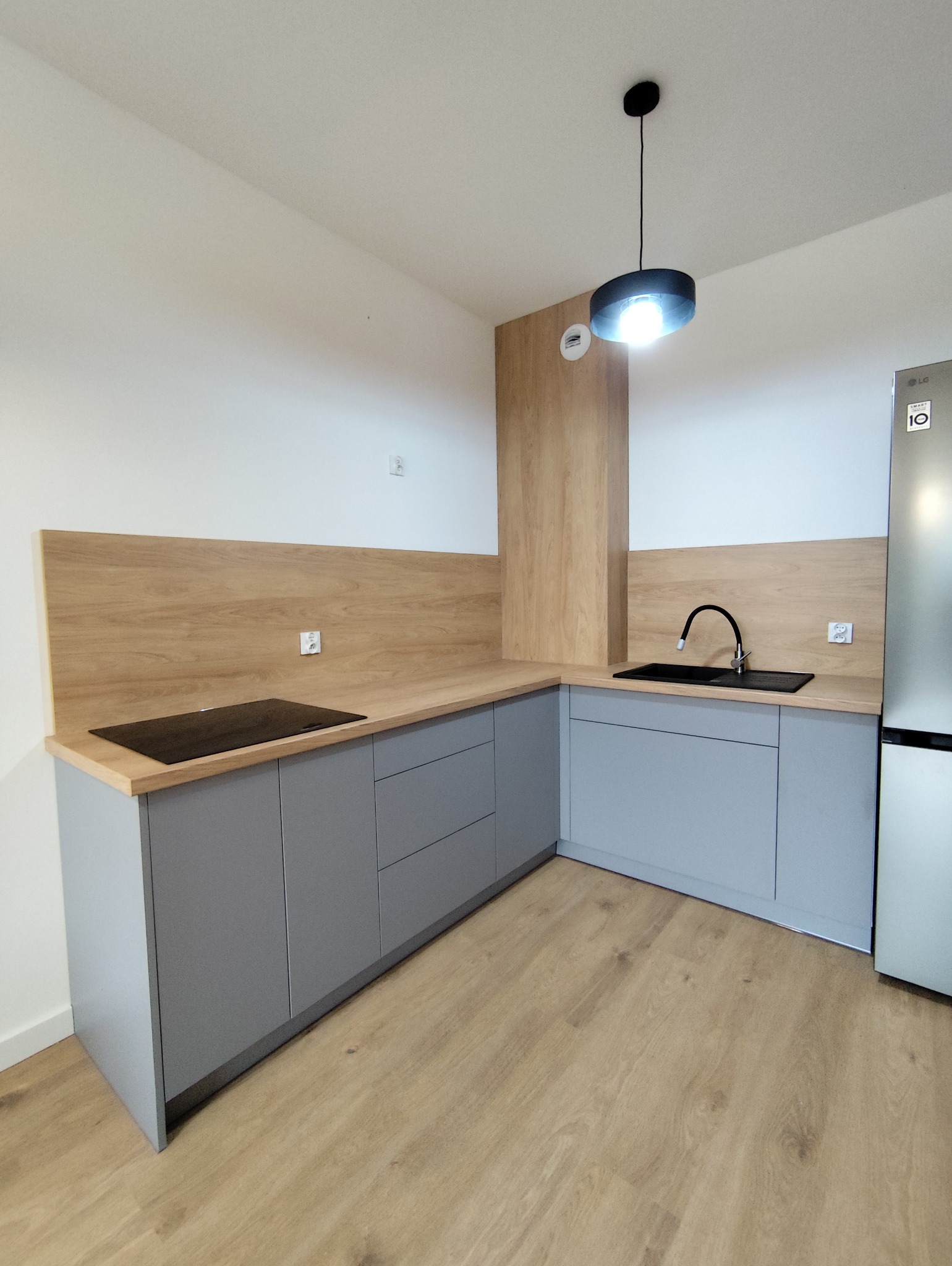 Szara zabudowa kuchenna z drewnianym blatem i panelem ściennym, czarny zlew i płyta indukcyjna, nowoczesna lampa sufitowa, jasna podłoga. Minimalistyczny design.