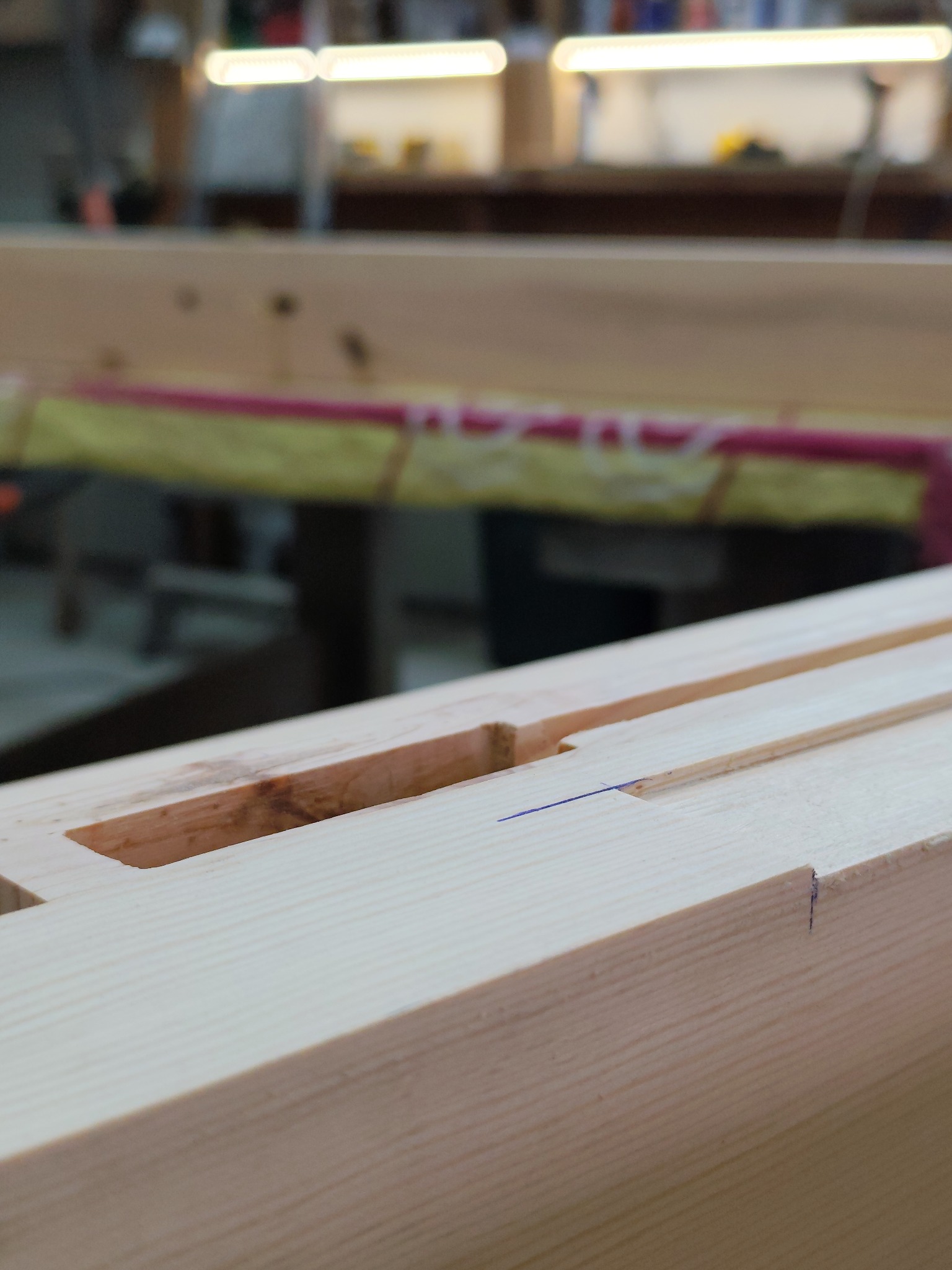 Detail of a light wood beam with a precisely cut mortise joint, marked with a blue line, suggesting meticulous woodworking in a workshop setting.