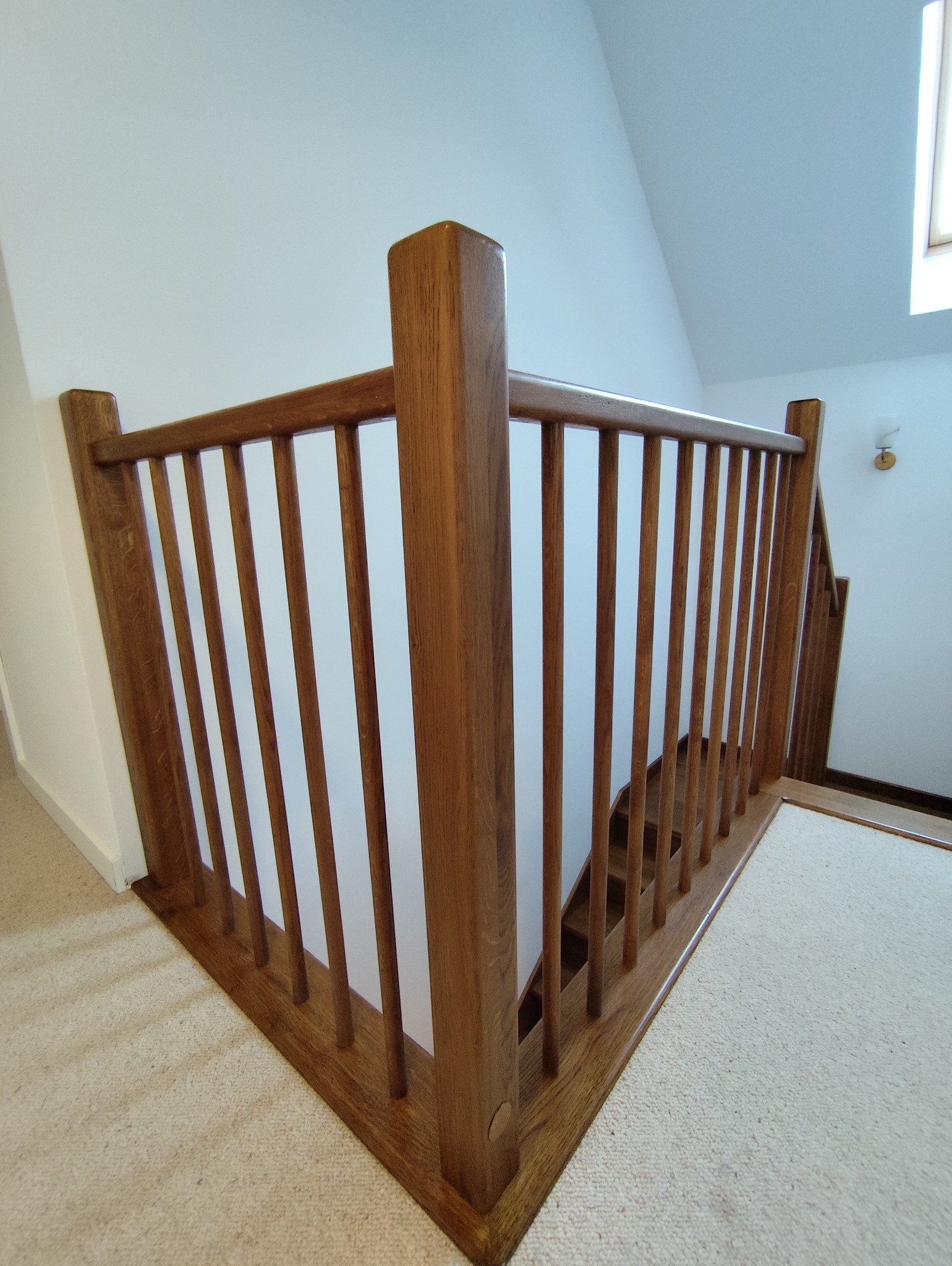 Drewniana balustrada schodowa z jasnego drewna, widok z dołu, na tle białych ścian i beżowej wykładziny. Element wykończenia wnętrza.