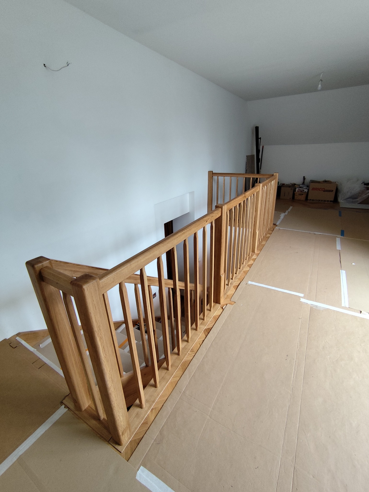 Drewniana balustrada schodowa z jasnego drewna, widok z góry, zabezpieczona podłoga kartonem, białe ściany, surowe wykończenie, wnętrze domu w trakcie prac.