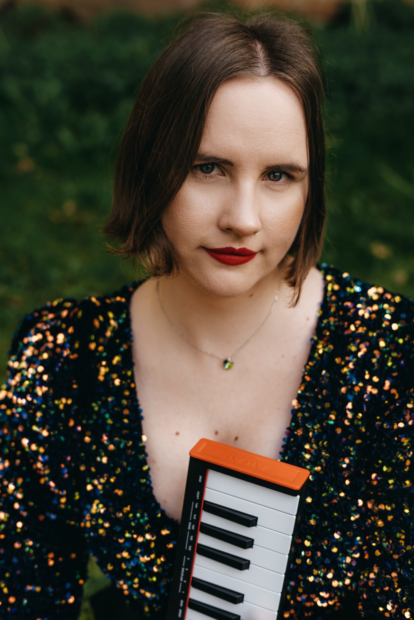 Portret kobiety z krótkimi włosami i czerwonymi ustami, trzymającej mały keyboard na tle zieleni. Ubrana w błyszczącą sukienkę.