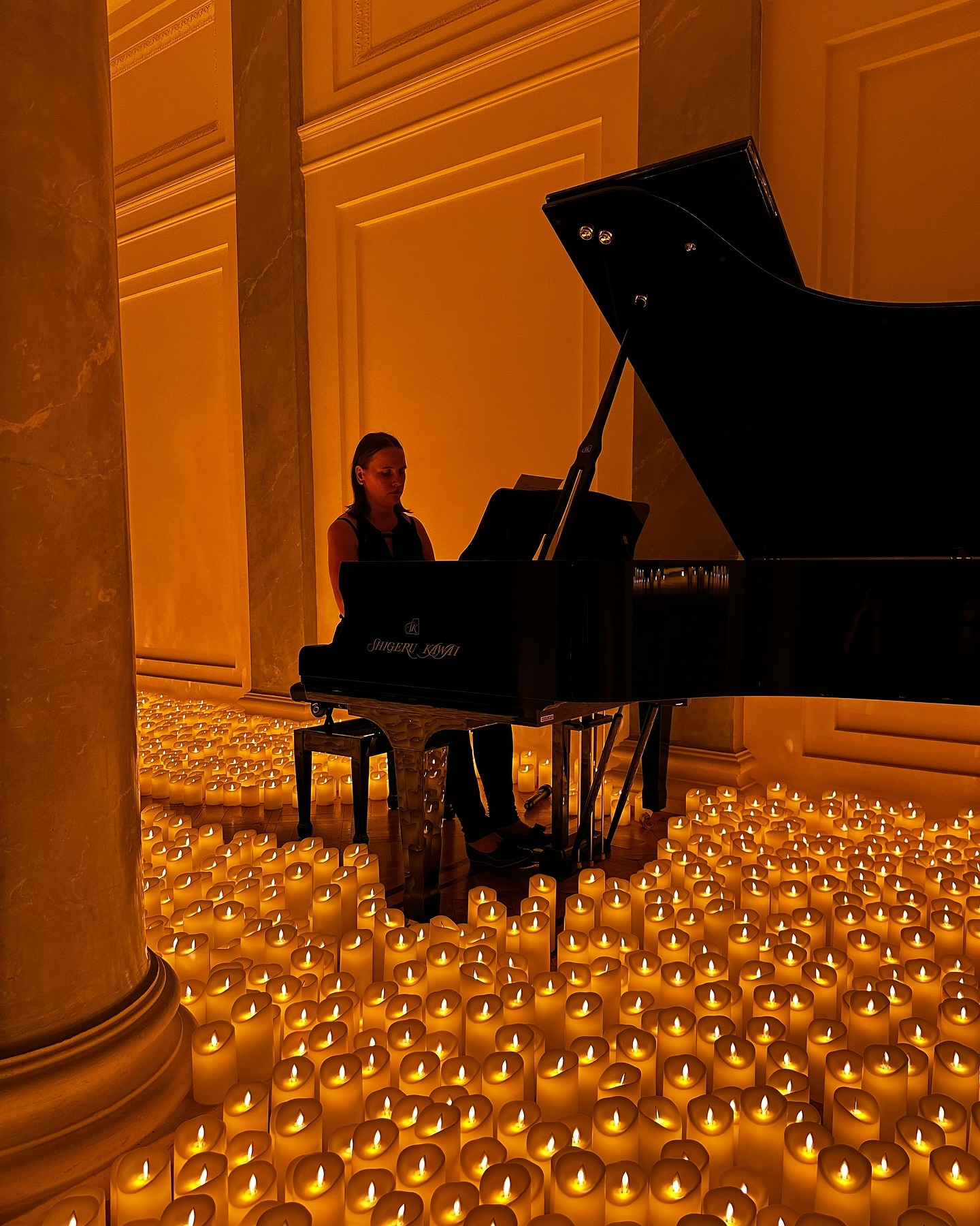Pianistka gra na fortepianie Shigeru Kawai w sali oświetlonej setkami świec, tworząc intymną atmosferę koncertu. Kolumny i ciepłe światło podkreślają elegancję.