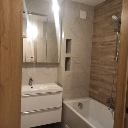 Oleksandr Shyrochenko - Nowoczesna łazienka po remoncie: biała umywalka z szafką, wanna, marmurowe i drewnopodobne płytki, wnęki w ścianie. Widok zza drewnianej ościeżnicy.