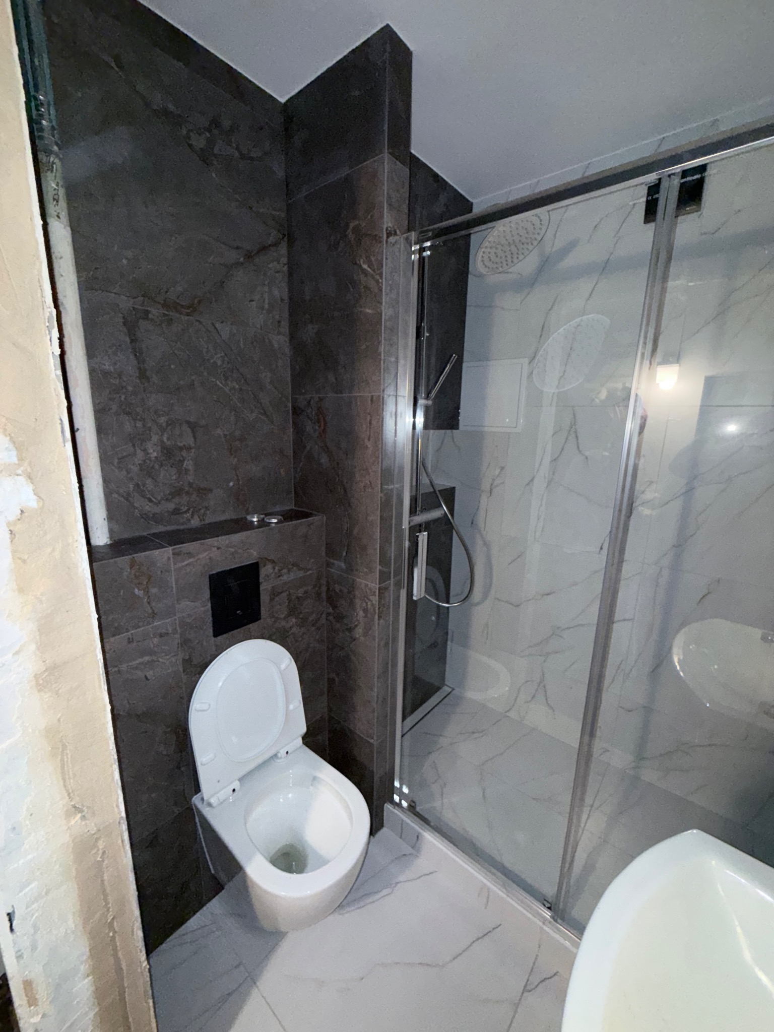 Nowoczesna łazienka w trakcie wykańczania: szare płytki imitujące marmur, biała ceramika sanitarna, kabina prysznicowa ze szklanymi drzwiami i fragment ściany przed wykończeniem.