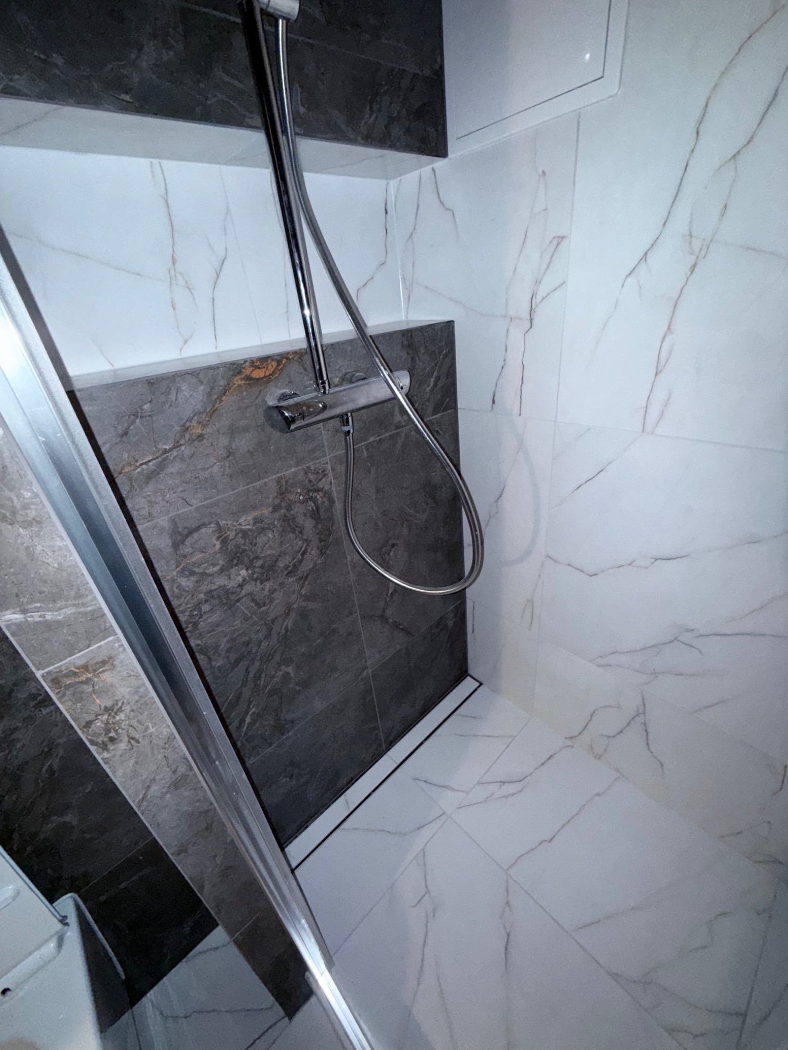 Nowoczesna kabina prysznicowa z szarymi i białymi płytkami imitującymi marmur, chromowaną armaturą i odpływem liniowym. Minimalistyczny design.