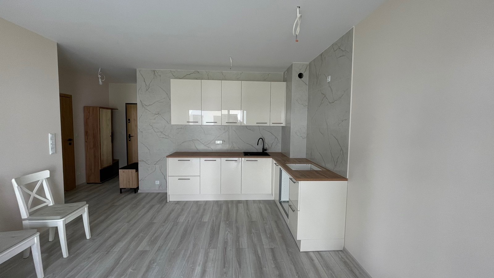 Jasne wnętrze po remoncie: biała zabudowa kuchenna z drewnianym blatem, szare płytki imitujące marmur na ścianie, panele podłogowe. Widok na przedpokój z szafą.