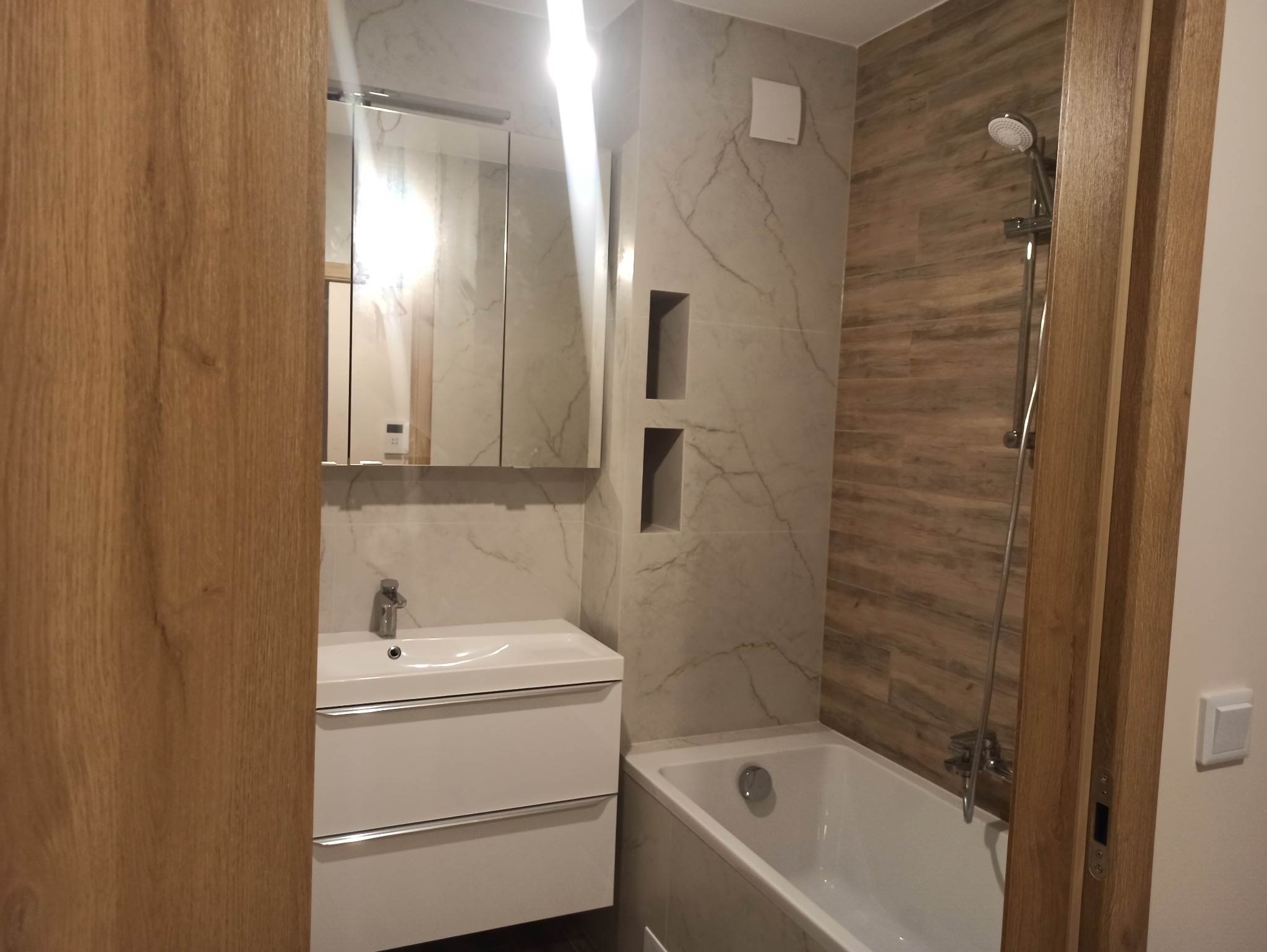 Nowoczesna łazienka po remoncie: biała umywalka z szafką, wanna, marmurowe i drewnopodobne płytki, wnęki w ścianie. Widok zza drewnianej ościeżnicy.