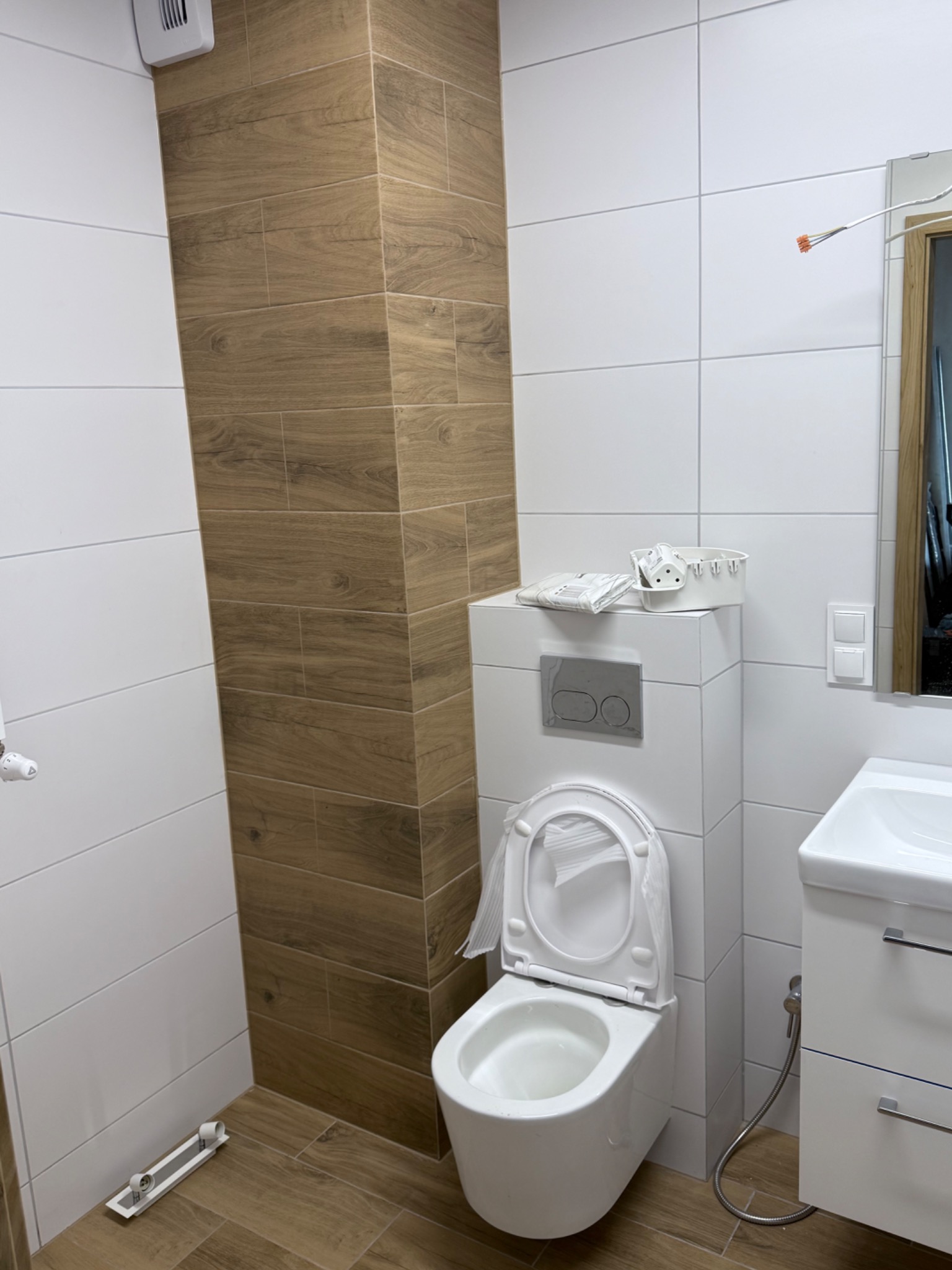 Nowoczesna łazienka po remoncie: białe i drewnopodobne płytki, wisząca toaleta z elementami montażowymi, umywalka z szafką. Widoczne detale wykończeniowe.