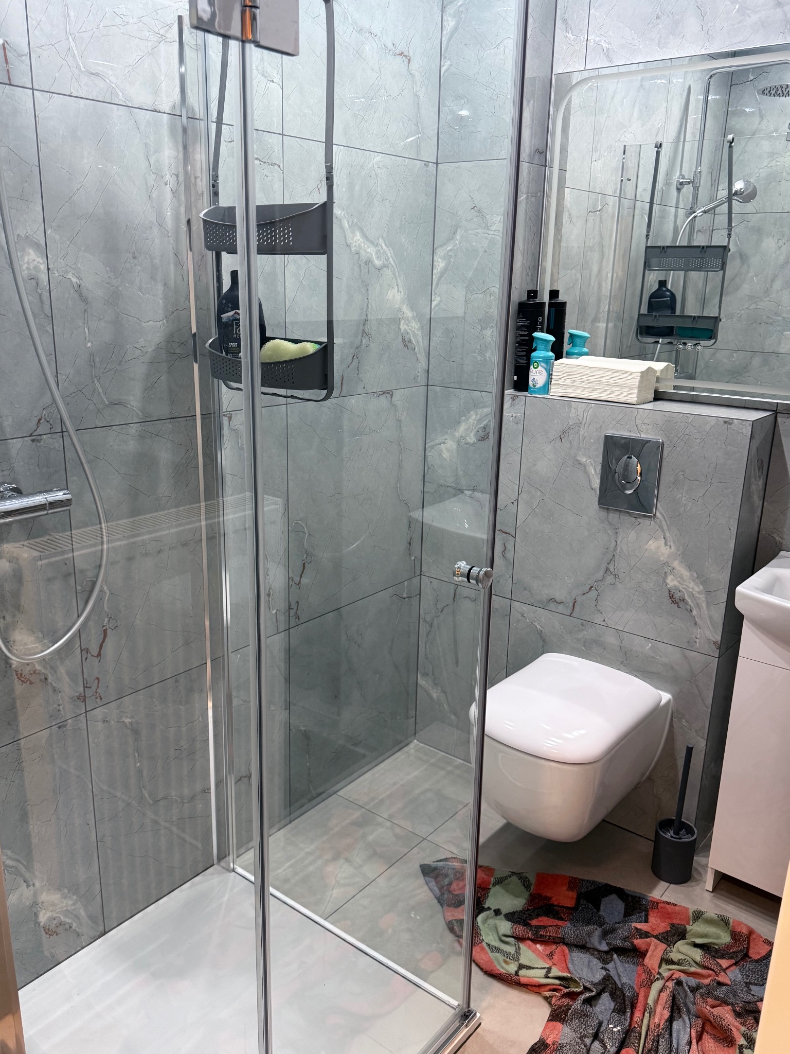 Wykończona łazienka z kabiną prysznicową, toaletą podwieszaną i szarymi płytkami imitującymi marmur. Widoczny fragment szafki z umywalką i akcesoria łazienkowe.