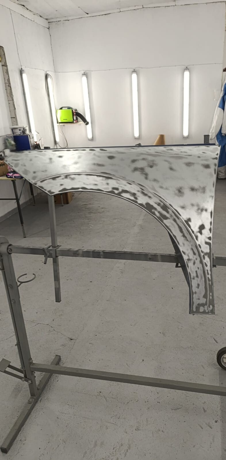 Przygotowany do lakierowania błotnik samochodowy na metalowym stojaku w warsztacie. Widoczne ślady szlifowania i przygotowania podłoża. Oświetlenie warsztatowe.