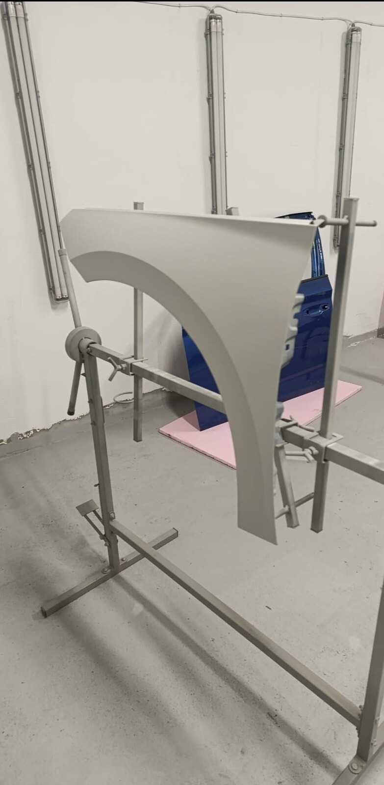 Pomalowany na szaro błotnik samochodowy zawieszony na metalowym stojaku w warsztacie, w tle niebieskie drzwi samochodowe i lampy oświetleniowe.