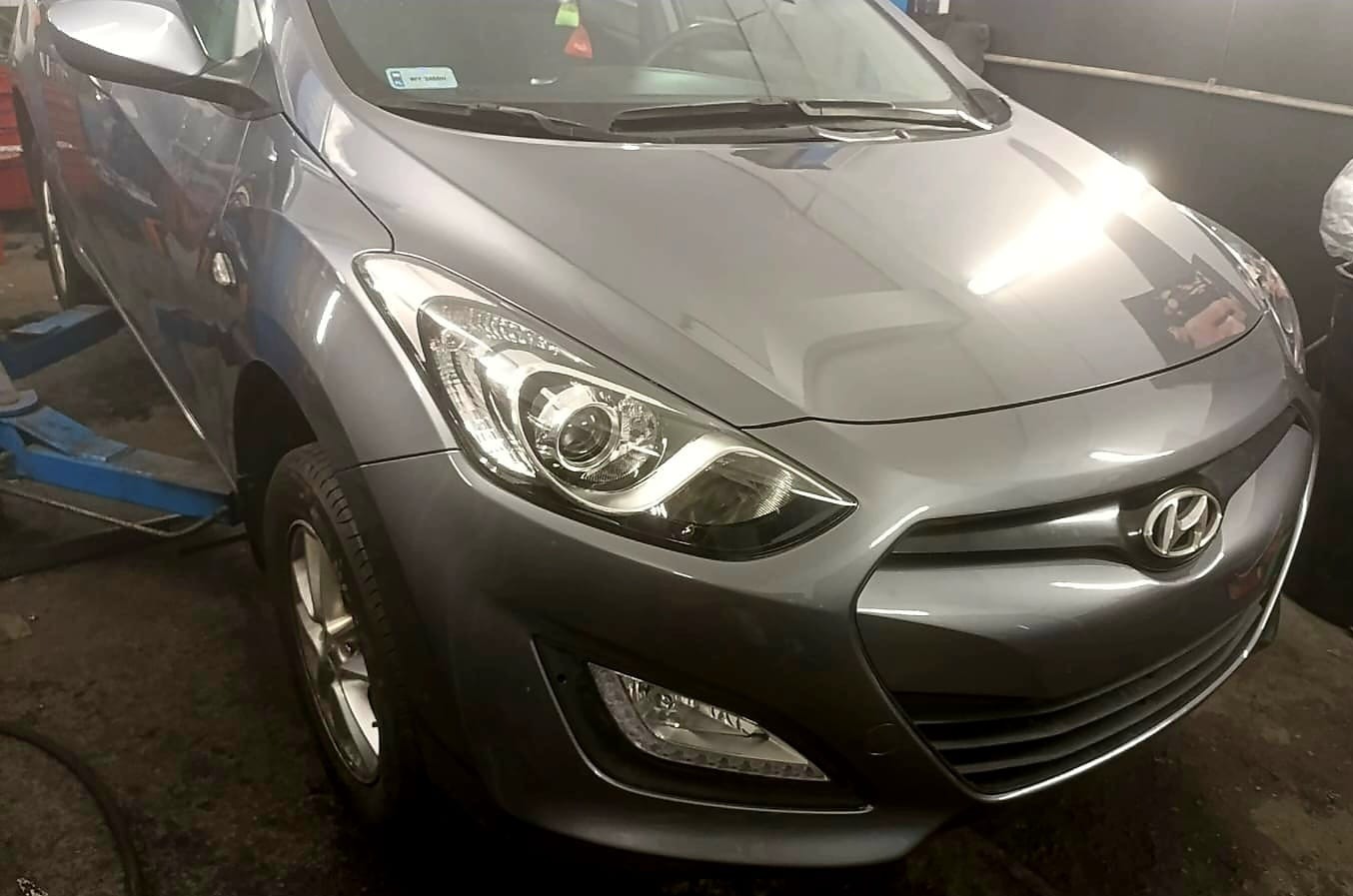 Szary Hyundai na podnośniku w warsztacie, widok z przodu, reflektor włączony. Widoczne ślady użytkowania na karoserii.