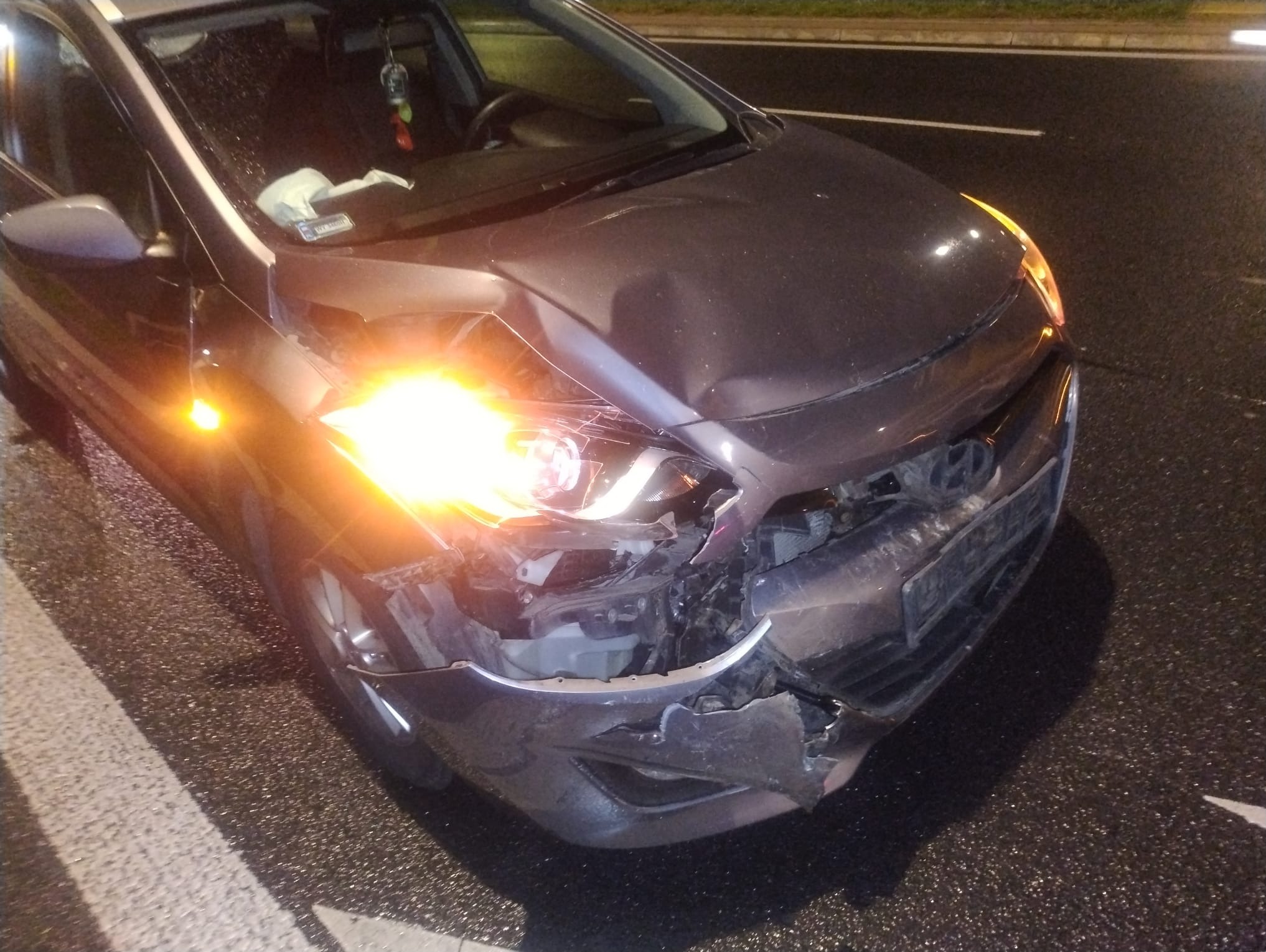 Uszkodzony przód szarego samochodu po kolizji w Mościskach. Zniszczony zderzak, reflektor i maska. Widoczne ślady uderzenia. Awaryjne światła włączone.
